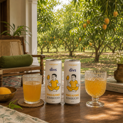 Two cans of divs Mango Kombucha are displayed on a wooden table in a patio setting, with two glasses of the golden beverage and a lush outdoor mango orchard in the background.

