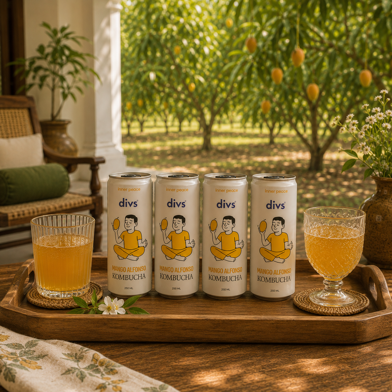 Four cans of divs Kombucha are arranged on a wooden tray next to two glasses of the amber drink, set in a rustic porch with an outdoor orchard in the background.